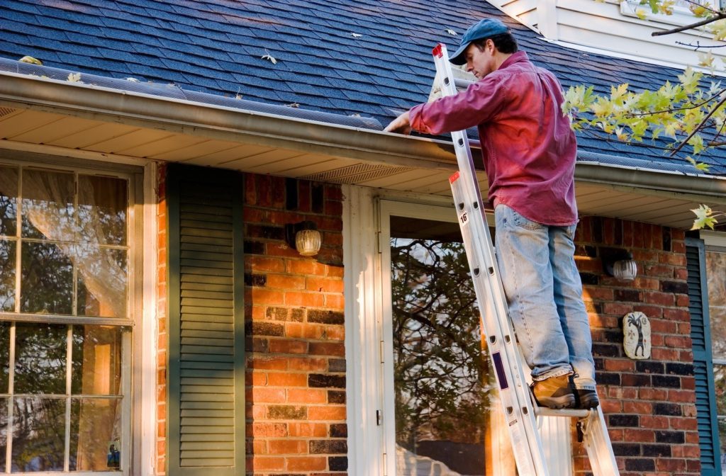 Man standing 5 feet up a ladder leaning against a house roof looking while he looks into a gutter on an autumn day