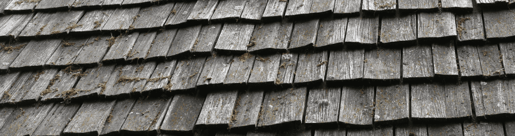 An image of an outdated wood shake roof, featuring rustic and traditional wooden roof tiles of an old house. The image is stretched and cropped across the banner.